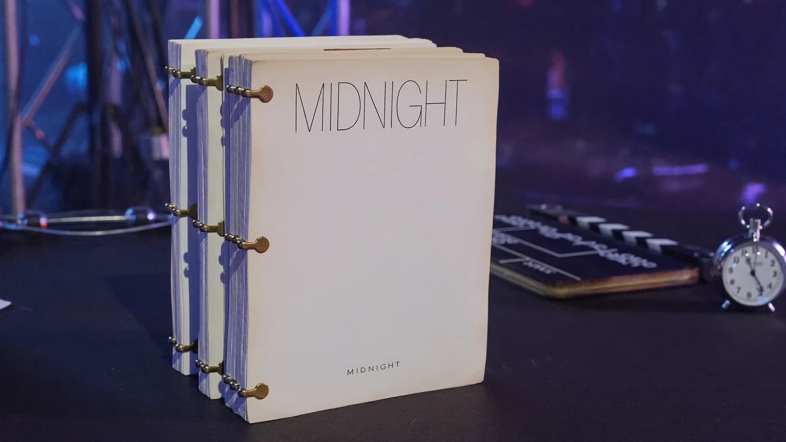 A thick feature screenplay titled Midnight standing beside a clapperboard and stopwatch on a dark film-set desk, cinematic still