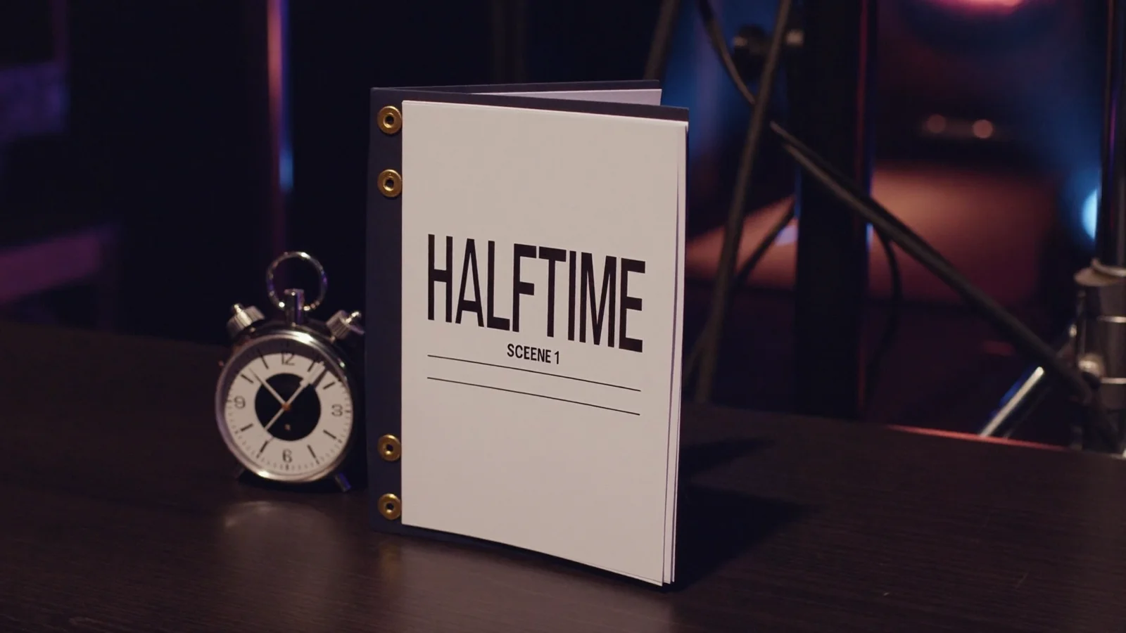A screenplay titled Halftime standing beside an analog stopwatch on a dark studio table, cinematic still