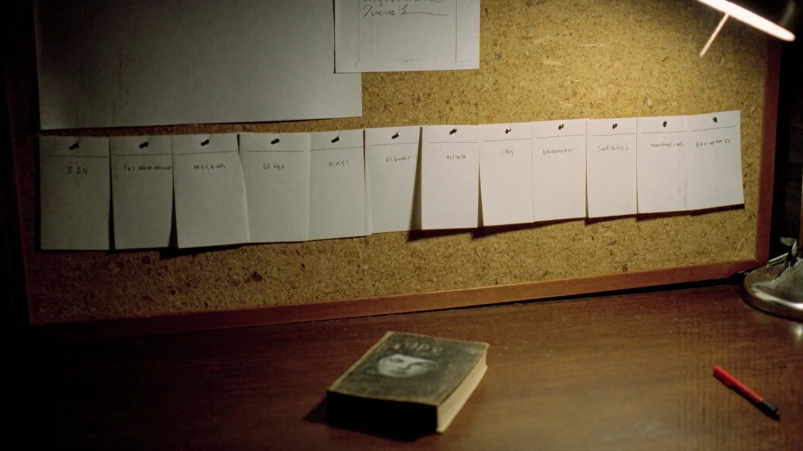 A screenwriter's corkboard with a row of blank index cards pinned under a warm desk lamp, a paperback and a red pen resting below, cinematic still