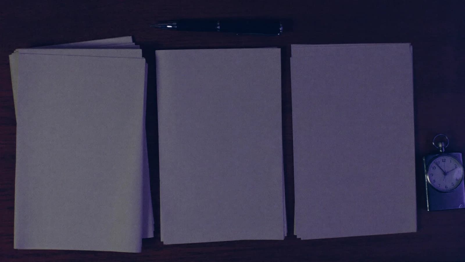 Three stacks of blank screenplay paper of different heights beside a pocket stopwatch and pen, overhead cinematic still