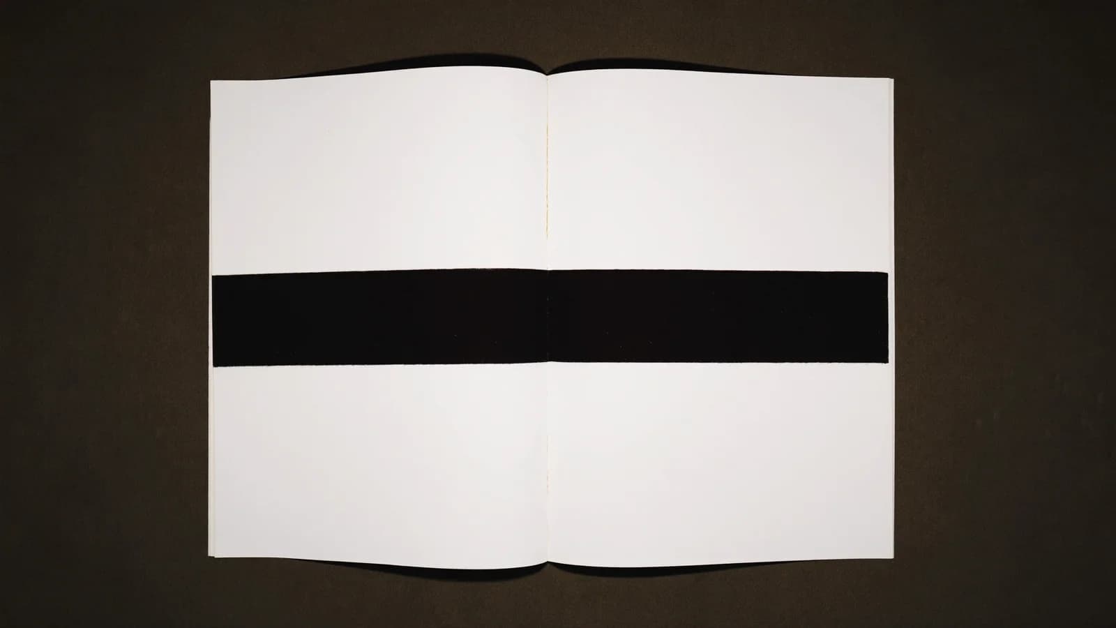 An open blank manuscript with a solid black redaction bar across both pages where identifying text would go, overhead cinematic still
