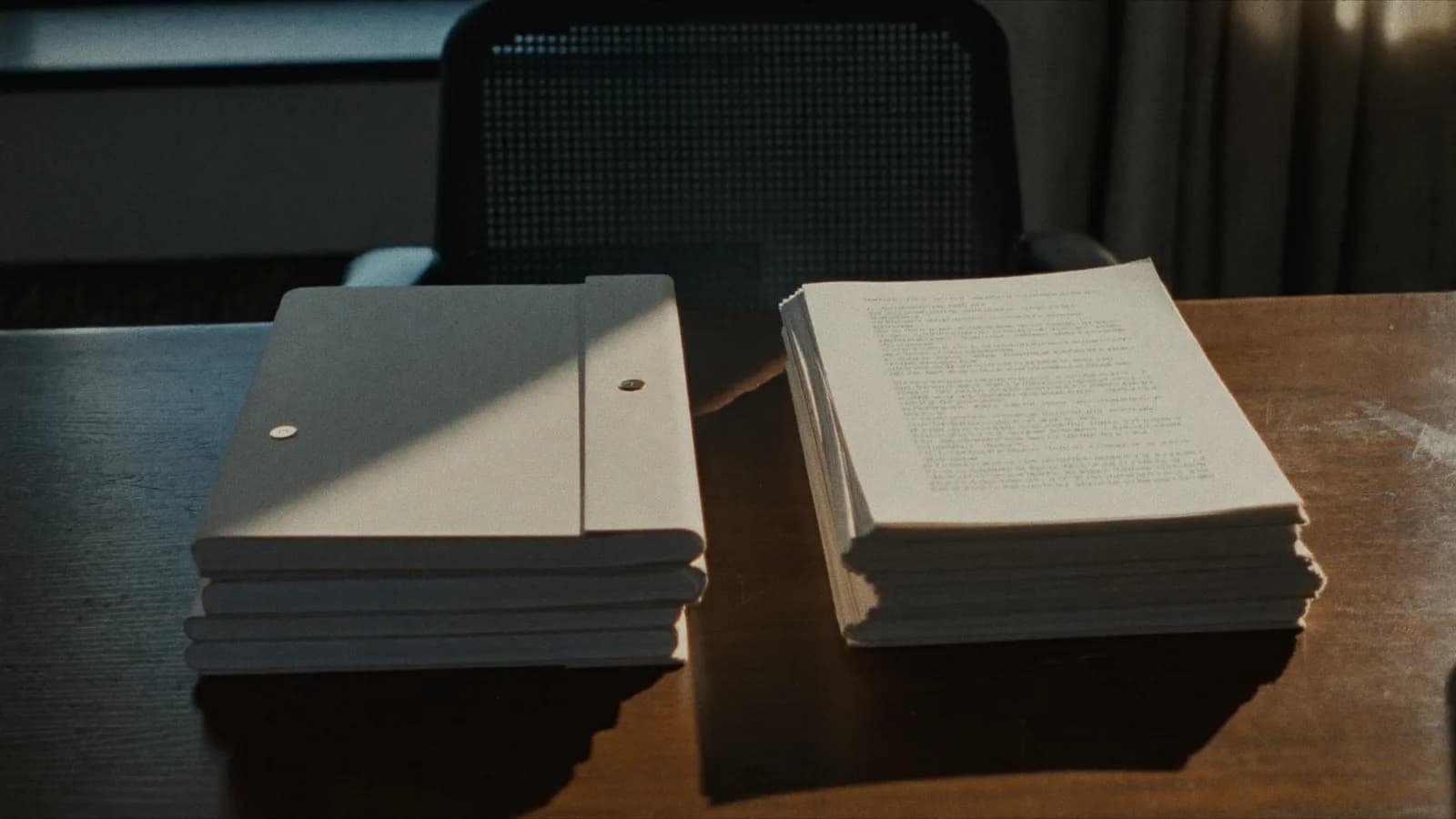 Two stacks of screenplays on a script reader's desk, one bound in plain card stock and one open to a typed first page, late afternoon light, cinematic still