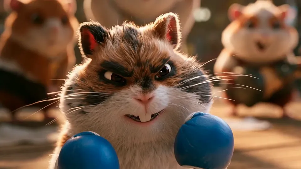 A calico hamster with dark fur around its eyes grins behind two blue boxing gloves