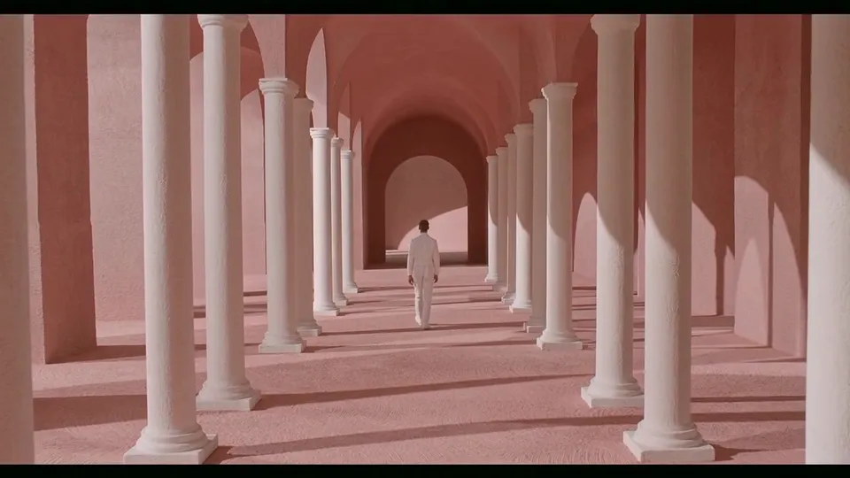 A small figure in white walking down a long pink colonnade in hard sunlight