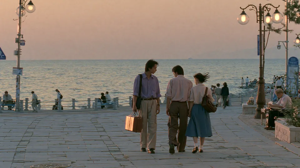 Three young people walk along a seaside promenade at sunset, one holding a small suitcase
