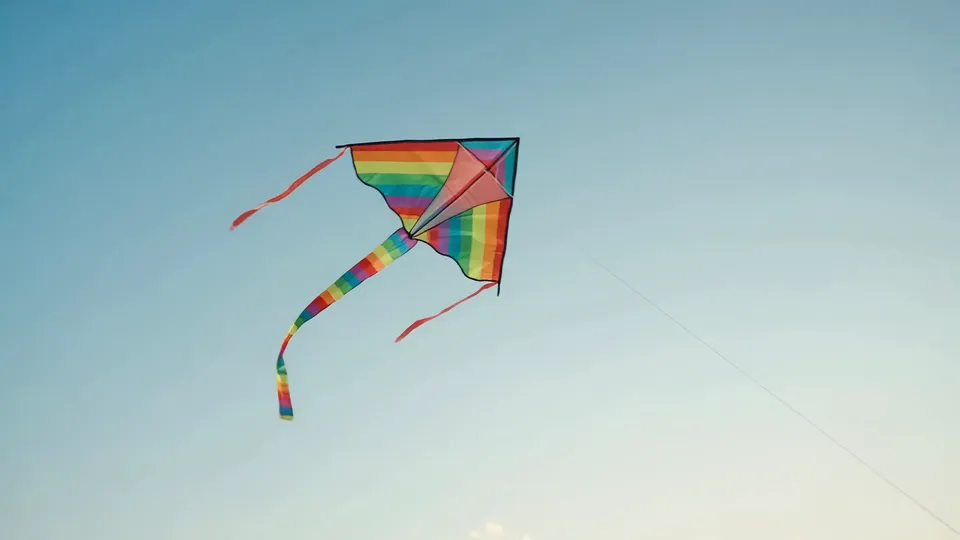 A rainbow kite with a long tail against a pale blue sky