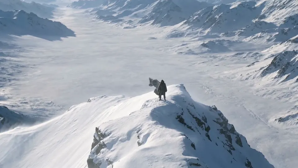 Aerial view of a lone cloaked traveller on a narrow snowy ridge above a white valley
