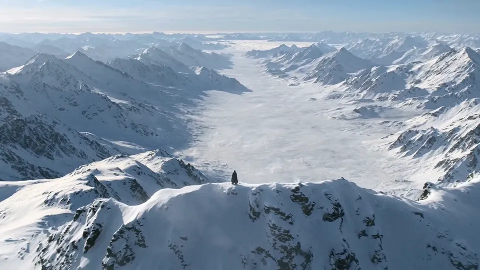 A tiny figure alone on a snowy ridge above a glacier valley that runs to the horizon