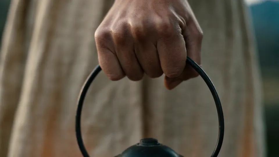A hand tightly gripping the iron ring handle of a lantern against a linen skirt