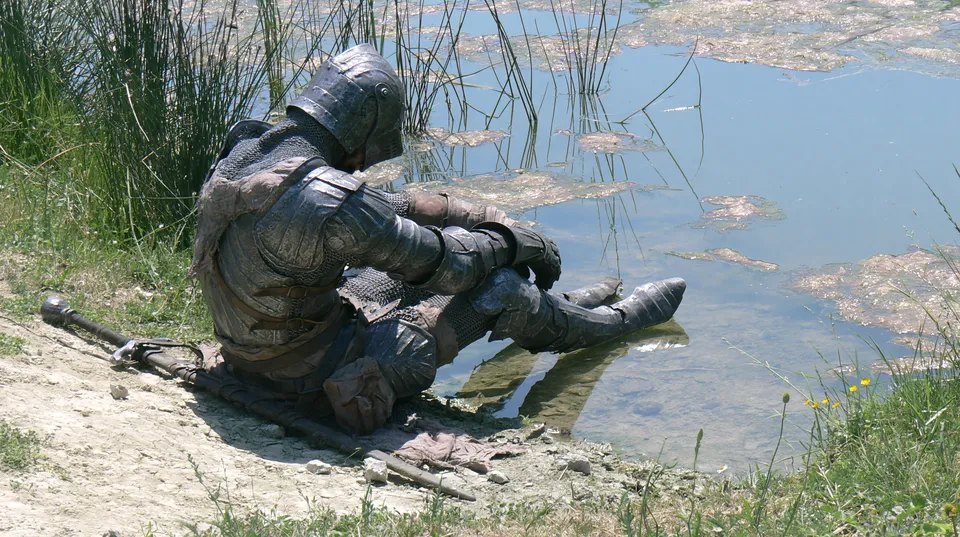 A knight in plate armour and chain mail sits at the edge of a pond, a mace lying in the dirt beside him