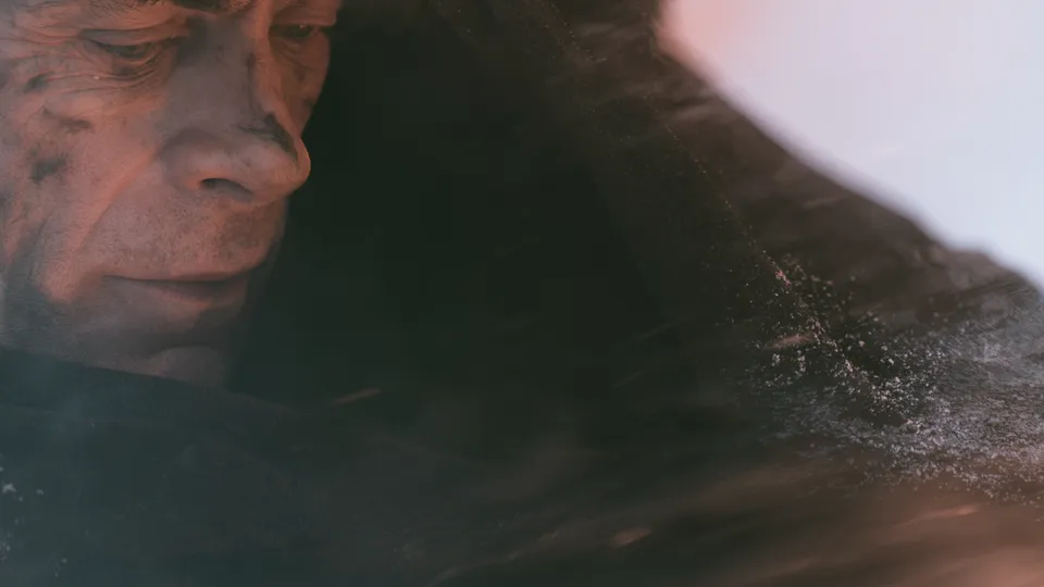 Close-up of an old man with a dusty, scarred face inside a hood, sand blowing past