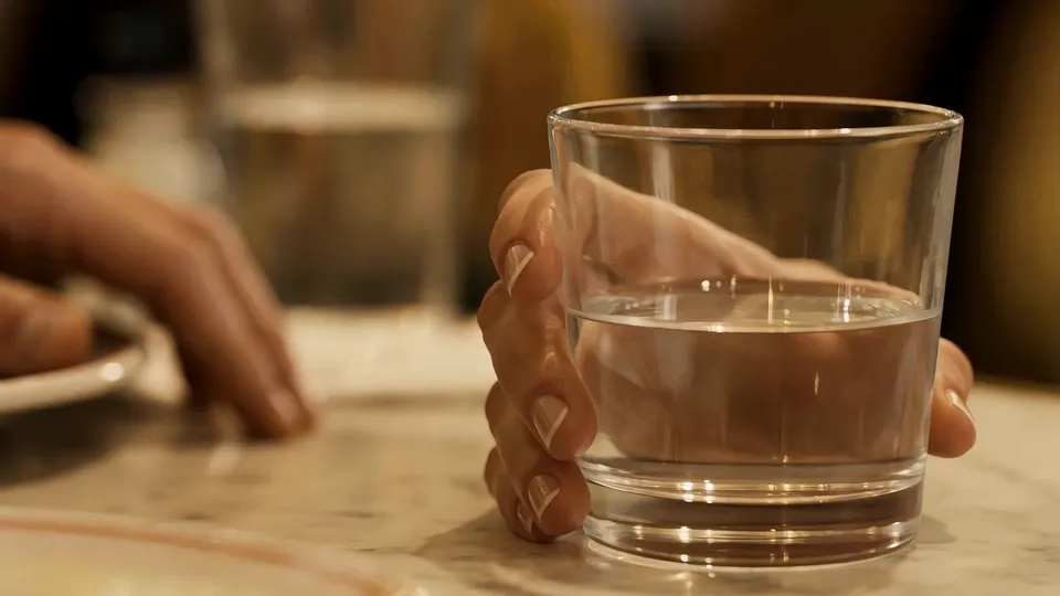 Primo piano di dita intorno a un bicchiere d'acqua su un tavolino di marmo, un'altra mano appoggiata lì vicino